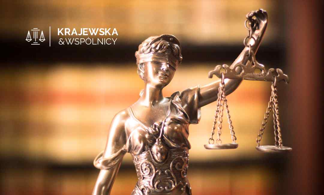 Metalowa statuetka Temidy, bogini sprawiedliwości z zasłoniętymi oczami i wagą szalkową, stoi na tle rozmytych grzbietów książek prawniczych