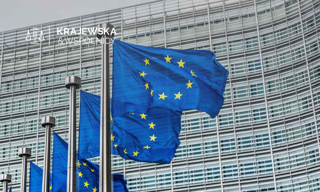 Flaga Unii Europejskiej z dwunastoma żółtymi gwiazdami na niebieskim tle powiewa na maszcie przed nowoczesnym, przeszklonym budynkiem