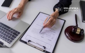 Na białym biurku widoczne są dłonie osoby używającej laptopa i wskazującej długopisem na dokument na podkładce, obok której leży drewniany młotek sędziego (gavel)