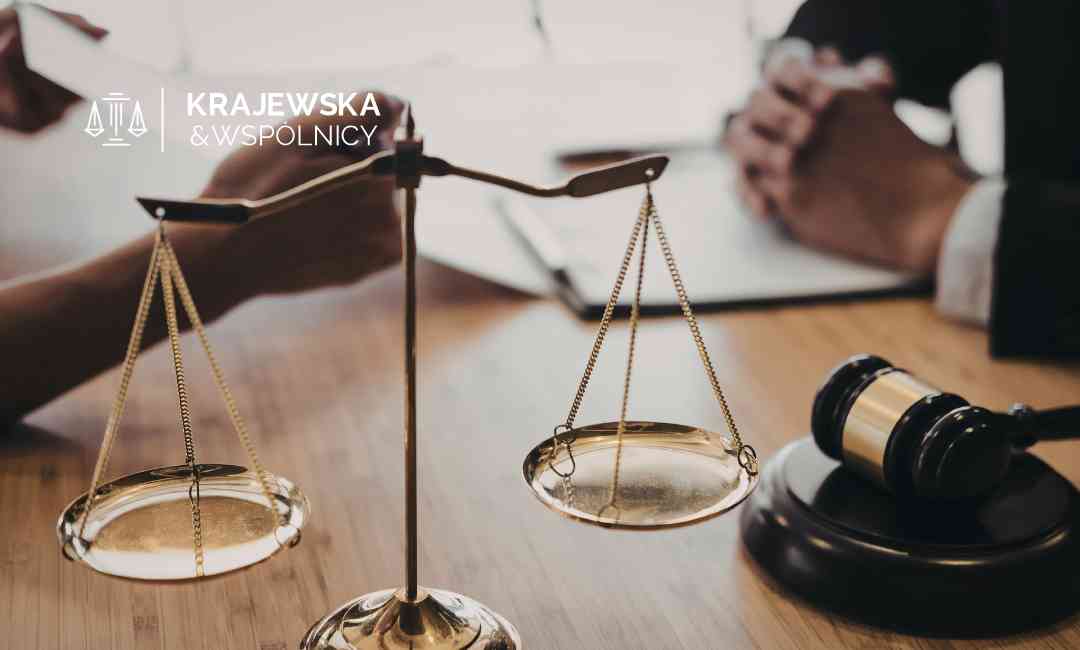 Złota waga sprawiedliwości oraz czarny młotek sędziowski na drewnianym biurku, w tle dłonie dwóch osób analizujących dokumenty podczas spotkania.