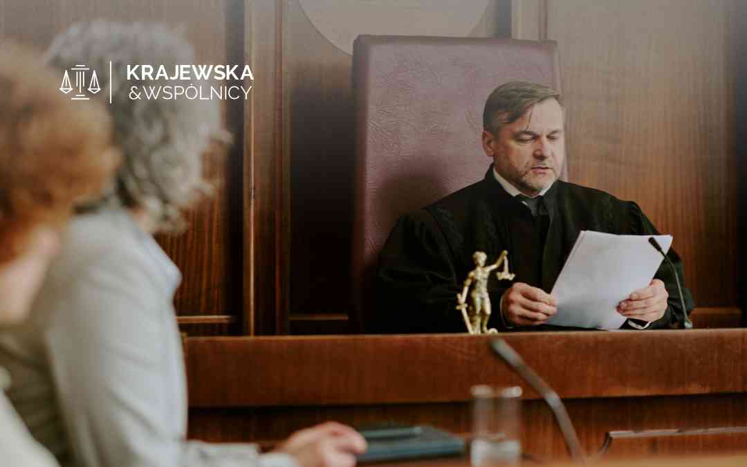 Sędzia w todze siedzący za wysokim stołem sędziowskim, czytający dokumenty na sali rozpraw. Przed nim widoczna mała figurka Temidy.