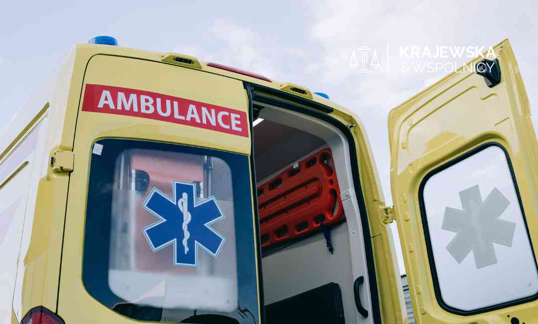 Widok z dołu na tył żółtego ambulansu z otwartymi drzwiami. Na oknie widoczna niebieska gwiazda życia, a powyżej czerwony napis „AMBULANCE”.