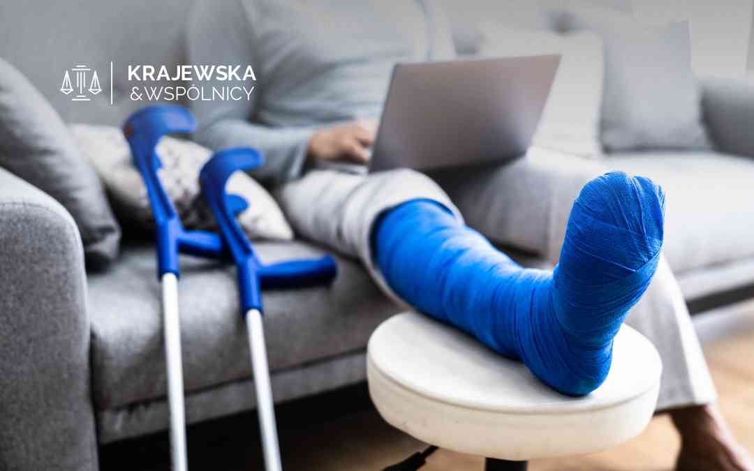 Osoba siedząca na szarej kanapie z nogą w błękitnym gipsie opartą na białym podnóżku. Obok oparte są niebieskie kule ortopedyczne, a na kolanach znajduje się laptop.