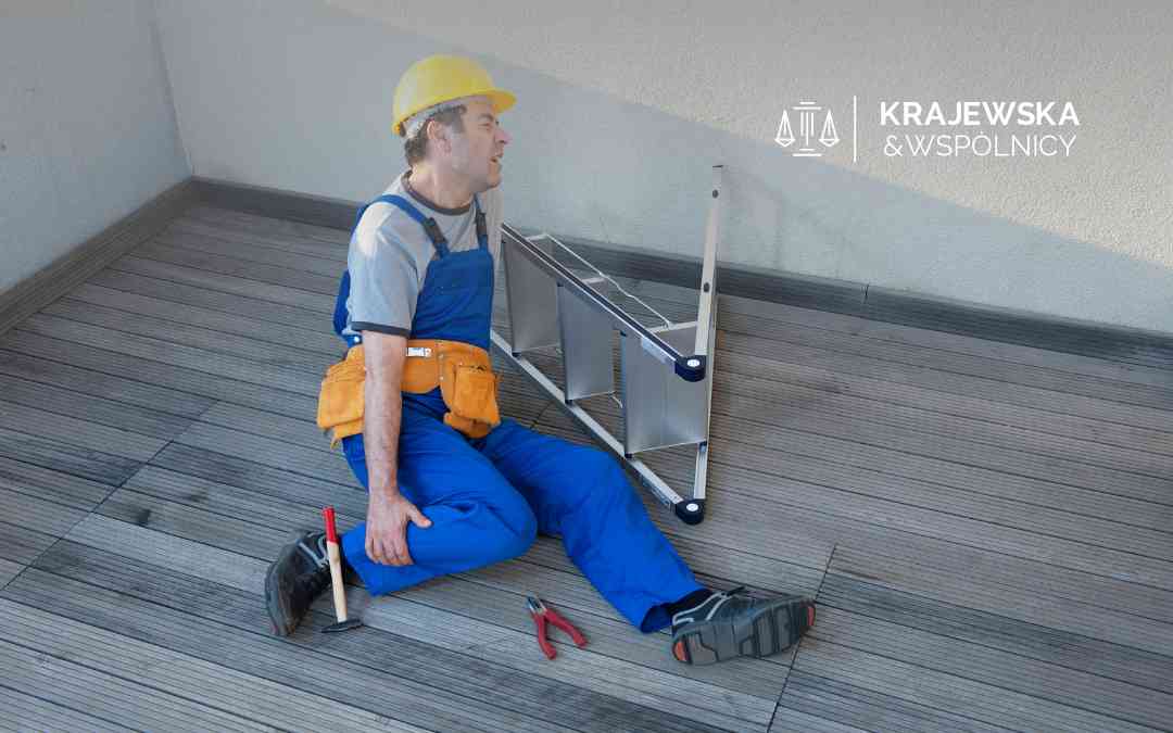 Monter w niebieskim kombinezonie siedzi na tarasie obok przewróconej drabiny i trzyma się za bolącą nogę. Przykład wypadku w pracy.