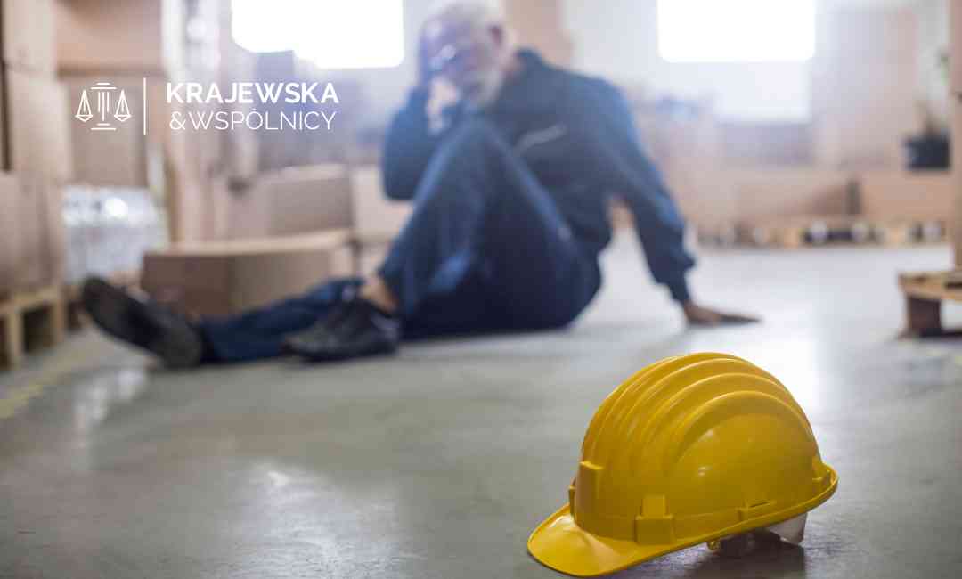 Pracownik siedzący na ziemi w magazynie trzyma się za głowę obok żółtego kasku – ilustracja wypadku przy pracy.