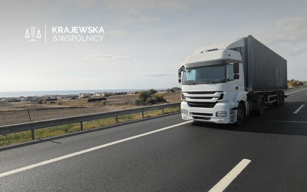 White semi-truck with a long container trailer driving on a coastal highway; distant buildings on the left and sea in the background.