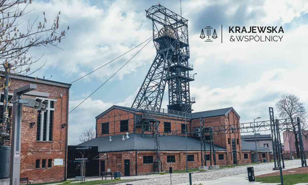 Brick industrial complex with a tall metal headframe, set under a partly cloudy sky; logo reads 'Krajewska & Wspólnicy' in the upper right.