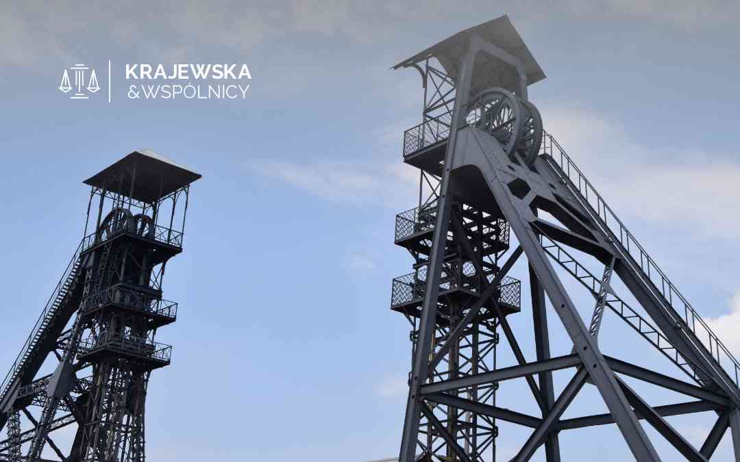 Two tall steel mining headframes with platforms and stairs stretch into a blue sky, with the watermark 'KRAJEWSKA & WSPÓLNICY' in the upper left.