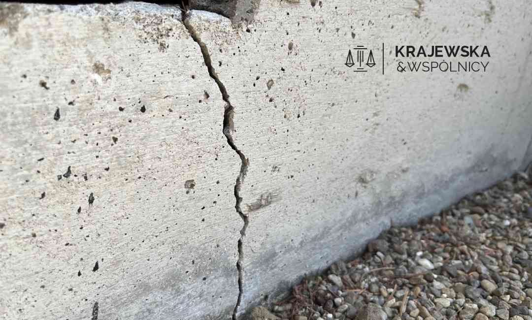 Cracked concrete wall with gravel at the base and a logo reading 'Kraje wska & Wspólnicy' in the top-right corner.