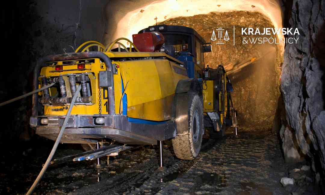 Underground mining vehicle (yellow excavator) inside a dim tunnel with rock walls and wet ground left by a work light glow.