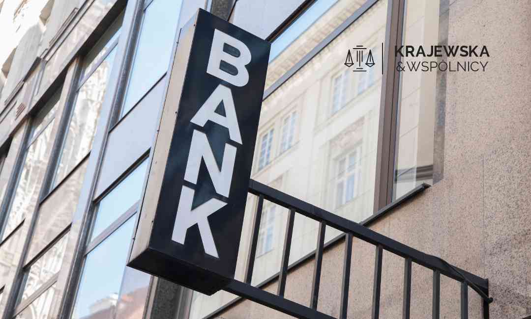 Pionowy czarny szyld z białym napisem BANK na tle szklanej fasady nowoczesnego wieżowca.